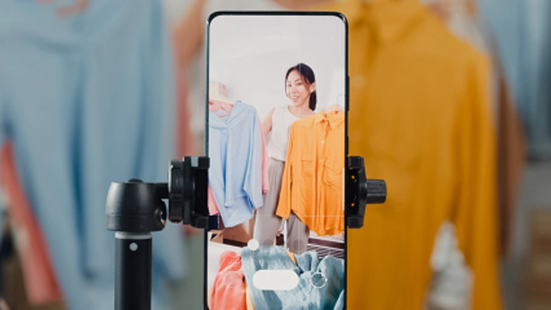 Person showcasing clothing items on a smartphone for social commerce livestream shopping with garments displayed in the background.