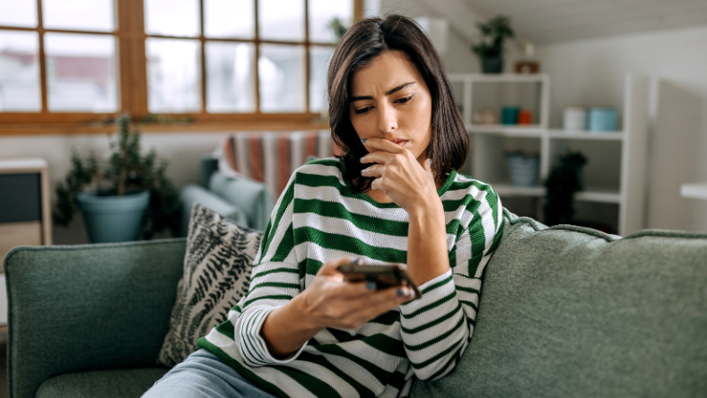 Woman sitting on couch very concerned while looking at mobile phone.