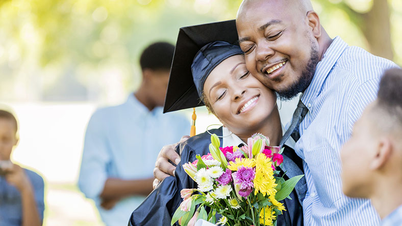 A woman in her graduation outfit hugging her husband and holding flowers
