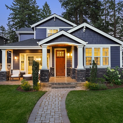 Exterior of a modern blue craftsman-style home with a covered front porch, stone columns, and warmly lit windows at dusk